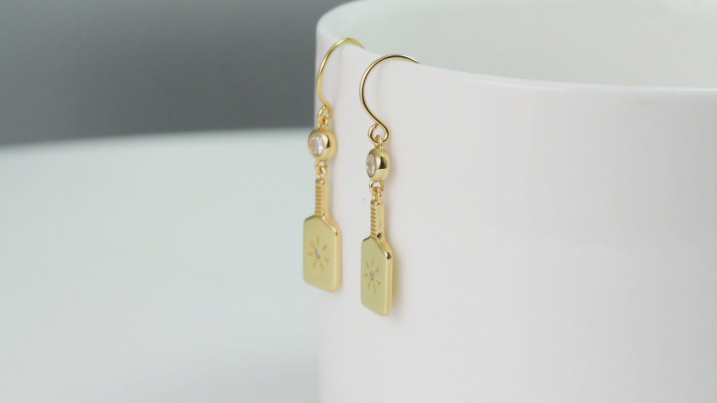 PickleBelle Paddle Drop Earrings in Gold. Features a dangling paddle below a sparkling crystal. Paddle even has a tiny stone and burst in the center reminiscent of a good hit on that sweet spot!These earrings dangle.